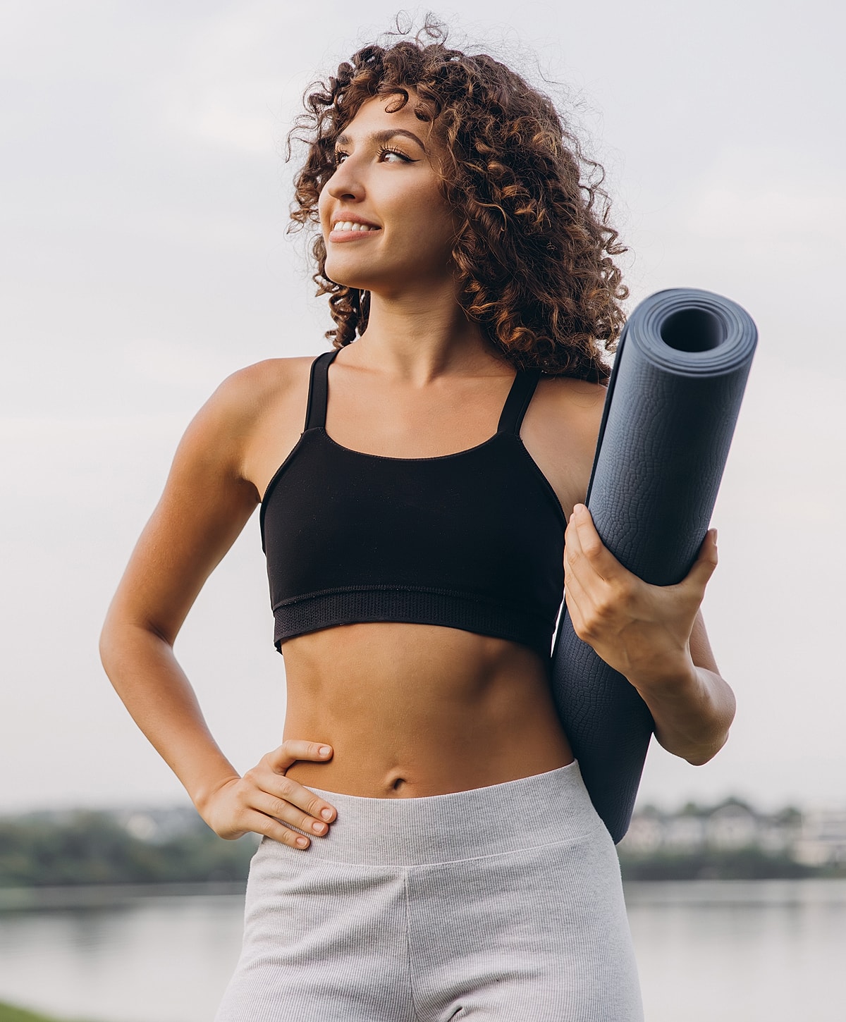 Woman holding yoga mat outdoors, smiling confidently.