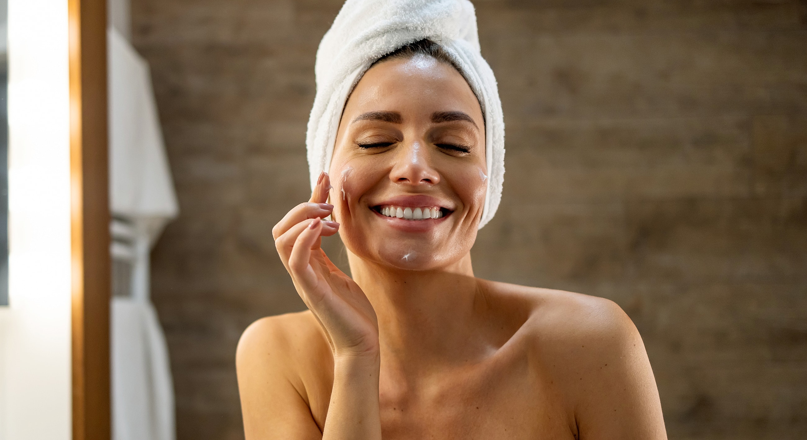 Woman applying skincare in bathroom with towel.